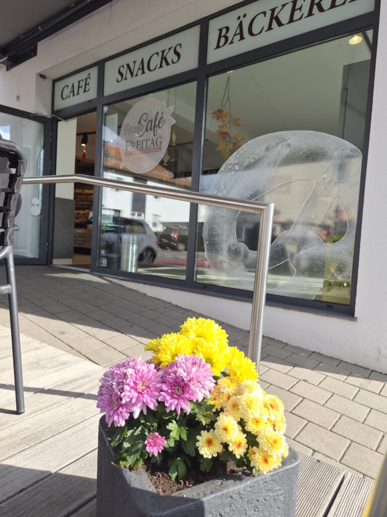 Eingang von Pascal’s Café Sindelfingen mit Café-Schriftzug, Brezelmotiv im Fenster und Blumen im Vordergrund